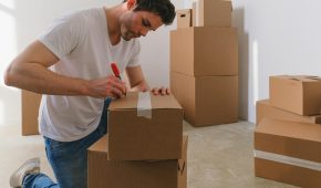 A man labels cardboard boxes for moving, kneeling indoors with natural light.